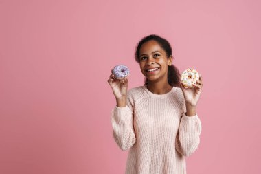 Brunette black girl laughing while posing with donuts isolated over pink background