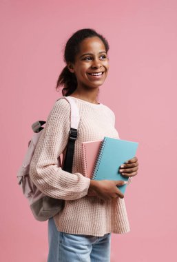 Black girl smiling while posing with exercise books and backpack isolated over pink background