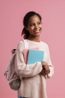 Black girl smiling while posing with exercise books and backpack isolated over pink background