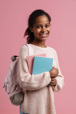 Black girl smiling while posing with exercise books and backpack isolated over pink background