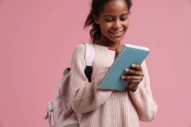 Black girl smiling while writing down notes in exercise book isolated over pink background