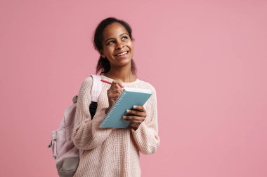 Black girl smiling while writing down notes in exercise book isolated over pink background