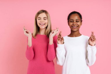 Multiracial two girls wearing sweaters holding fingers crossed isolated over pink wall