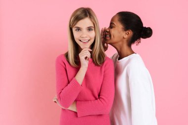 Black girl smiling while whispering secret to happy friend isolated over pink wall