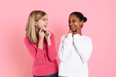 Multiracial two girls wearing sweaters pointing fingers at their smiles isolated over pink wall