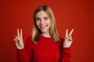 Blonde white girl laughing while showing peace sign isolated over red wall