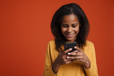 Brunette black girl smiling and using mobile phone isolated over red wall