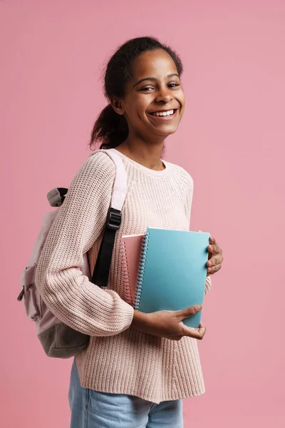 Black girl smiling while posing with exercise books and backpack isolated over pink background