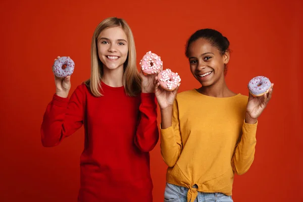Multiracial Two Girls Smiling While Posing Donuts Isolated Red Wall — стоковое фото