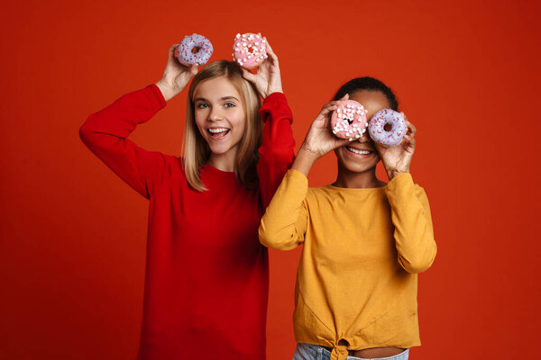 Multiracial two girls laughing while making fun with donuts isolated over red wall