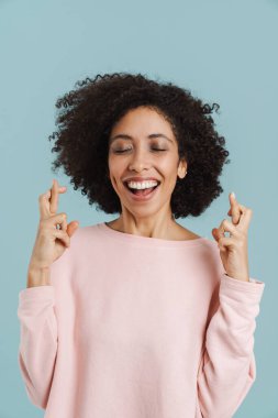 Black young woman smiling while holding fingers crossed for good luck isolated over blue background