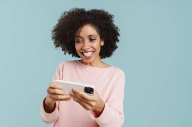 Young black woman smiling and playing online game on cellphone isolated over blue background