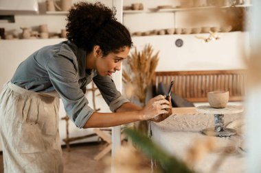 Young black ceramist woman taking photo on cellphone while working at her studio
