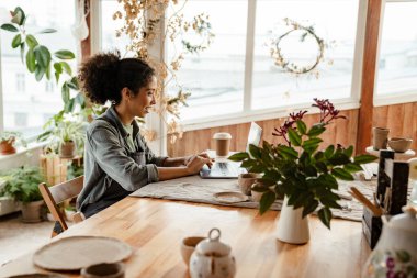 Young black ceramist woman smiling and using laptop while working in her studio