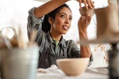 Young black ceramist woman wearing apron sculpting in clay at her workshop