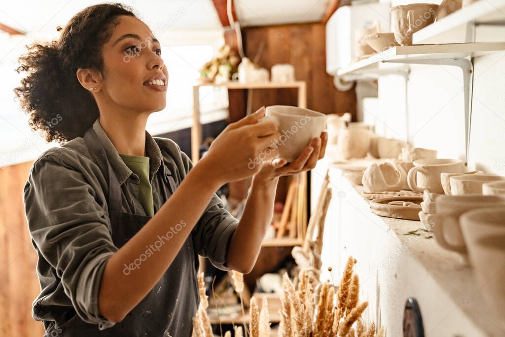 Young black ceramist woman wearing apron smiling while working in her studio