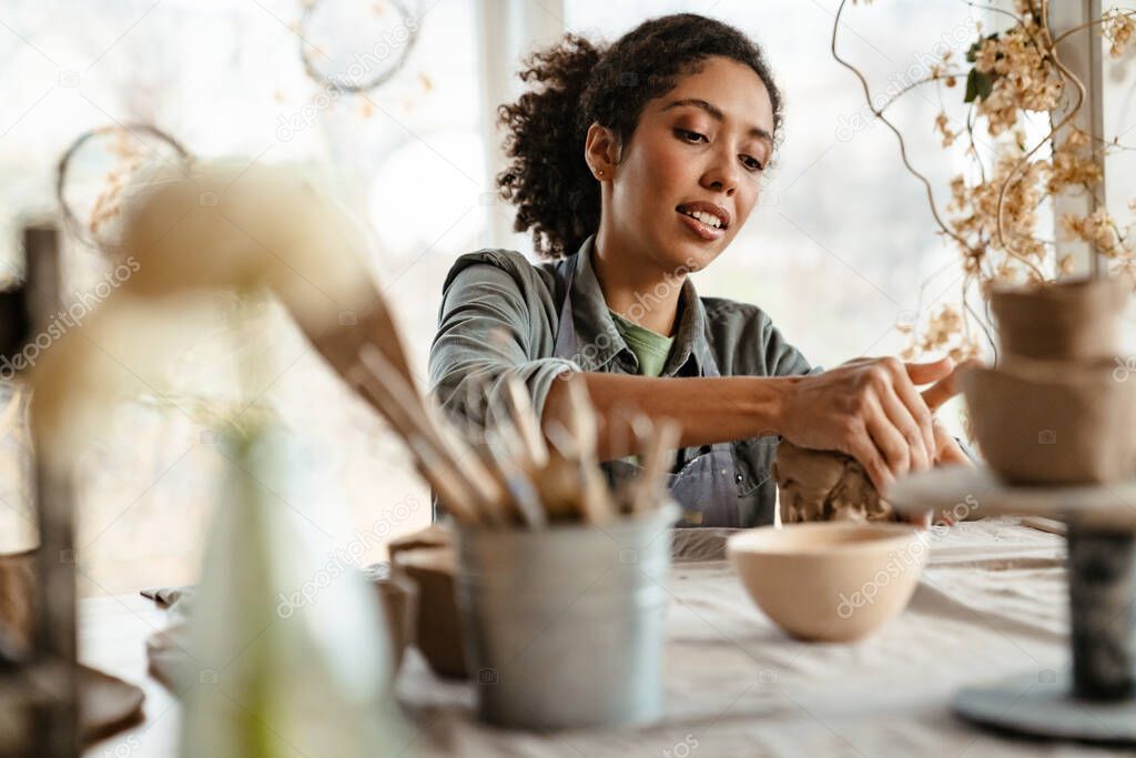 Young black ceramist woman wearing apron sculpting in clay at her workshop