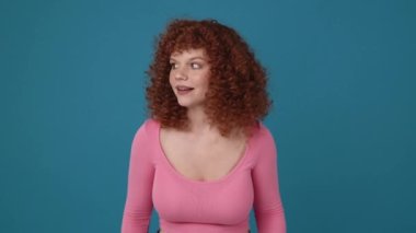 Happy curly haired redhead woman wearing pink shirt showing shh gesture at the camera in blue studio