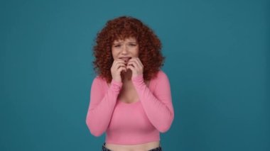 Scared curly haired redhead woman wearing pink shirt looking at the camera in blue studio