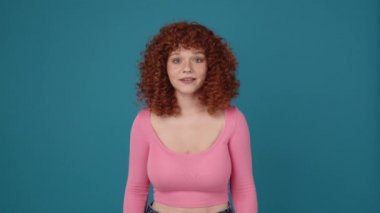 Cute curly haired redhead woman wearing pink shirt looking at the camera in blue studio