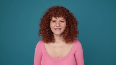 Handsome curly haired ginger woman wearing pink shirt hoping for something in blue studio