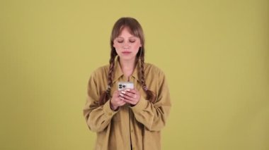Pensive blonde woman with pigtails thinking what to text on phone isolated over green background