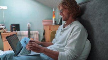 Smiling blond man wearing eyeglasses working on laptop while sitting on the bed at home indoors