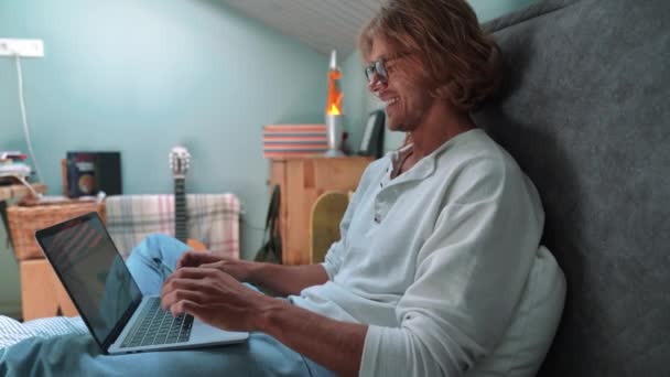 Smiling Blond Man Wearing Eyeglasses Working Laptop While Sitting Bed – Stock-video