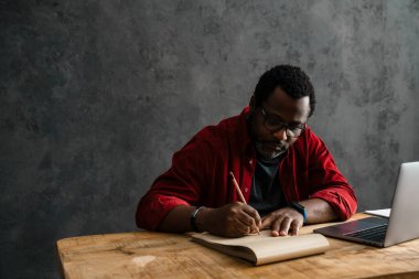 Black man in eyeglasses writing down notes while working with laptop indoors