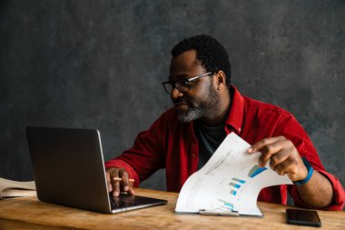 Black man in eyeglasses smiling while working with laptop and papers office
