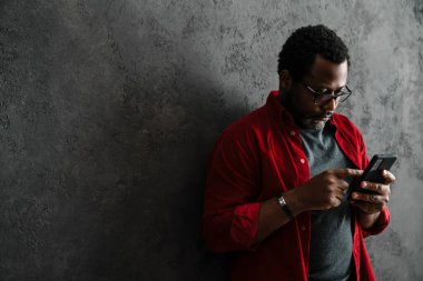 Black man in eyeglasses using mobile phone while leaning on concrete wall