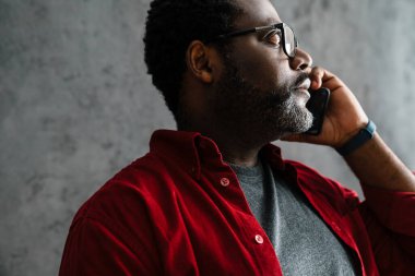 Black man in eyeglasses talking on cellphone while standing by concrete wall