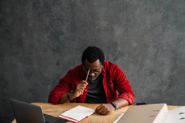 Black man in eyeglasses writing down notes while working with laptop indoors