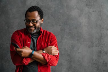 Black man in eyeglasses using cellphone while standing by concrete wall indoors