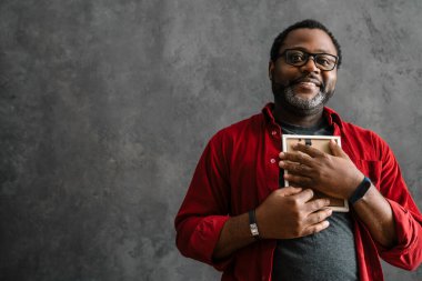 Black man in eyeglasses smiling and holding photo frame while standing by concrete wall indoors