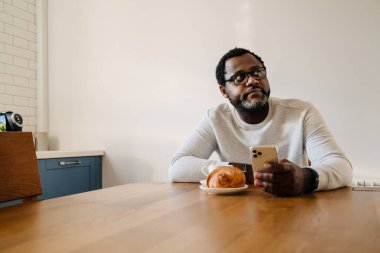 Black man wearing eyeglasses using mobile phone while having breakfast at home