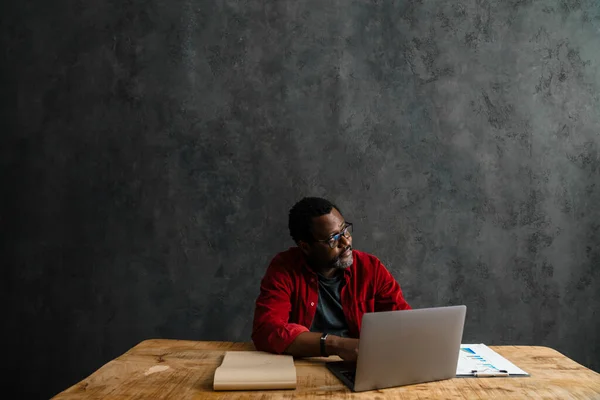 Black man in eyeglasses working with laptop while sitting at desk indoors