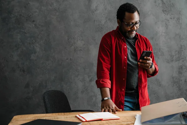 Black man in eyeglasses using cellphone while working at desk indoors