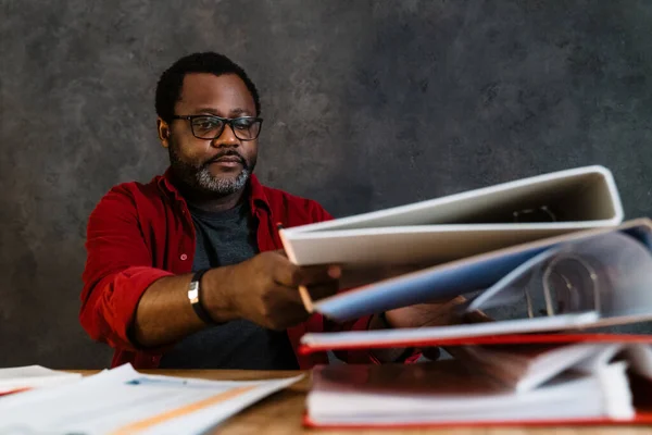 Black man in eyeglasses working with papers while sitting at desk indoors