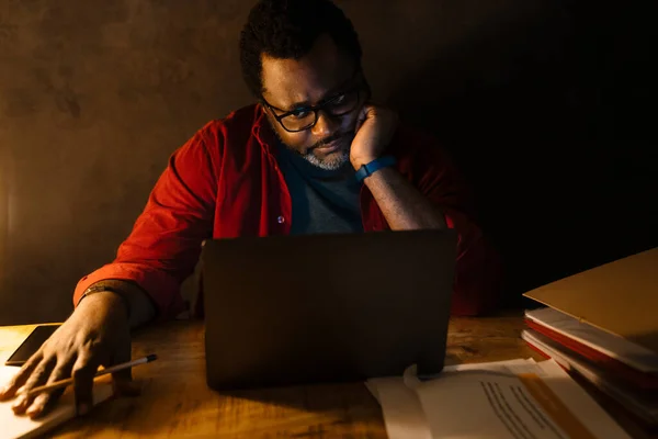 Black man in eyeglasses working with laptop while sitting at desk indoors