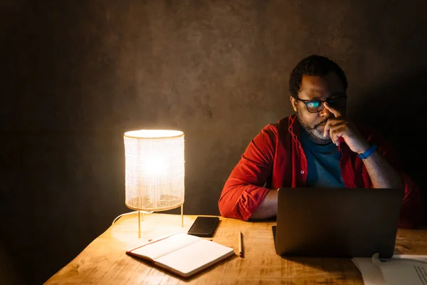 Black puzzled man in eyeglasses working with laptop while sitting at desk indoors