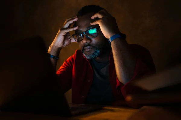 Black puzzled man in eyeglasses working with laptop while sitting at desk indoors