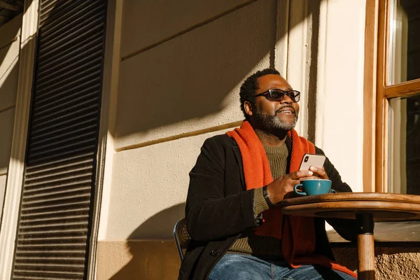 Black bearded man using cellphone and drinking coffee while sitting in cafe outdoors
