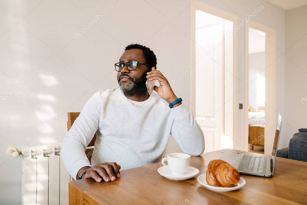 Black man wearing eyeglasses talking cellphone while having breakfast at home