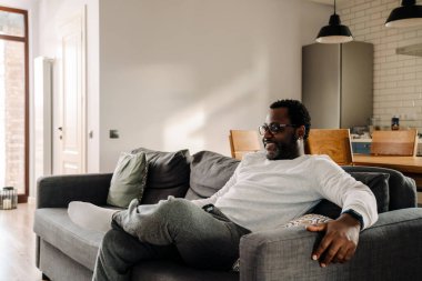 Black man wearing eyeglasses watching tv while resting on sofa at home