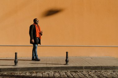 Black bearded man in sunglasses using cellphone while standing by wall outdoors