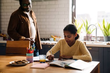 Black girl writing in exercise book while doing homework with her father at home