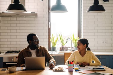 Black man working with laptop while his daughter doing homework at home