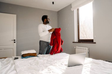 Black man using laptop while folding his clothes in bedroom at home