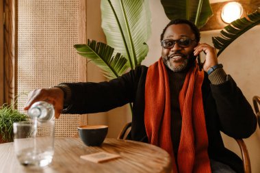 Black man talking on cellphone while drinking water in cafe indoors